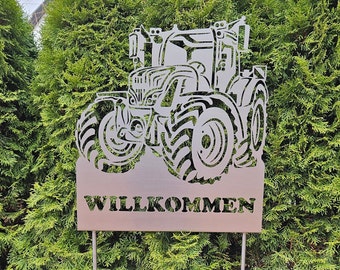 May include: A metal silhouette of a tractor with the word "WILLKOMMEN" on a sign below. The tractor is facing the right side of the image. The sign is brown and the tractor is silver.