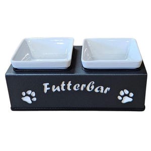 Könnte beinhalten: Ein schwarzes Futternapf-Set mit zwei weißen quadratischen Schalen. Die Basis ist schwarz mit dem Wort "FutterBar" in Weiß sowie Pfotenabdruck-Designs. Dieses Haustierzubehör ist für die Fütterung von Haustieren konzipiert.