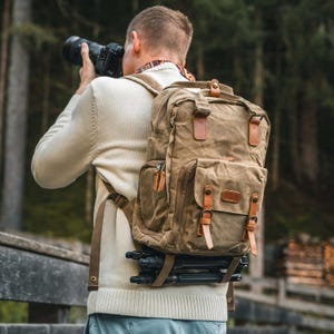 Puede incluir: Una mochila de lona color caqui con detalles y correas de cuero marrón. La mochila tiene múltiples bolsillos y un trípode adjunto. La persona lleva un suéter color crema y sostiene una cámara.