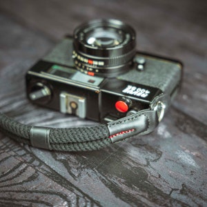 Può includere: Una macchina fotografica a pellicola nera da 35 mm con una tracolla in pelle nera. La macchina fotografica ha un pulsante rosso e il testo "ROCCOR" stampato sul lato.