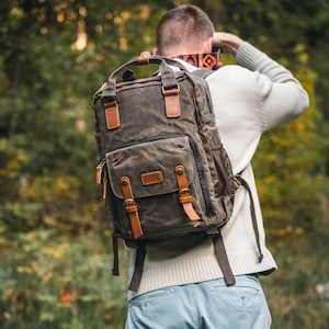 Può includere: Uno zaino in tela marrone con dettagli in pelle. Lo zaino ha più tasche, cinghie e fibbie. È dotato di una maniglia superiore e di una tasca laterale in rete. La borsa è indossata da una persona con un maglione color crema e pantaloni blu.