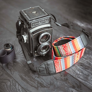 May include: A black Rolleicord camera with a colourful patterned camera strap. The camera has two lenses and a black leather strap. The camera is sitting on a dark wooden surface.