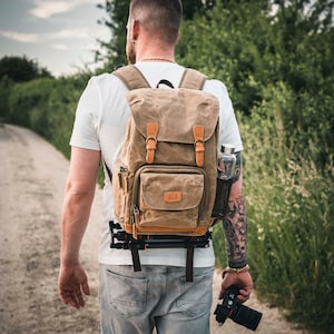 Peut inclure: Un sac à dos en toile beige avec des détails et des sangles en cuir marron. Le sac à dos comporte plusieurs poches et est fixé avec des boucles. Un trépied est attaché en bas. Une bouteille d'eau est dans une poche latérale.