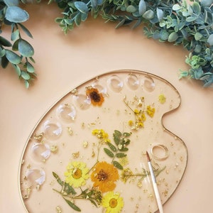 May include: A round, white, resin paint palette with a gold rim. The palette has 12 circular wells and is decorated with pressed flowers and gold leaf. A wooden paintbrush is lying next to the palette.