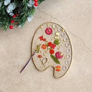 May include: A gold-rimmed artist's palette with a clear resin top. The resin contains pressed flowers and gold leaf. A paintbrush is lying next to the palette.