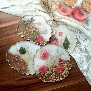 May include: Four round resin coasters with gold foil and pressed flowers. The coasters are white with a clear resin top and have a scalloped edge.