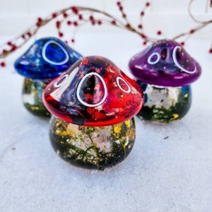 May include: Three resin mushroom ornaments with different colored tops. The mushrooms are filled with dried flowers and glitter. The tops are blue, red, and purple.