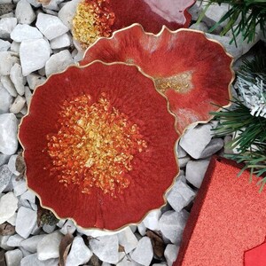 May include: Two red resin coasters with gold flecks and a wavy edge. The coasters are on a bed of white rocks.
