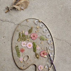 May include: A gold-rimmed, white ceramic palette with pressed flowers and gold leaf accents. The palette is on a grey concrete surface with a paintbrush lying next to it.