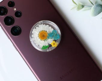 May include: A burgundy smartphone with a clear phone grip featuring pressed flowers. The phone grip is attached to the back of the phone.