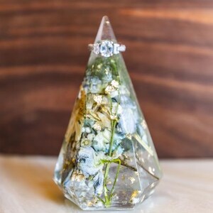 May include: A clear, geometric ring holder with a silver ring on top. The holder is filled with dried blue and white flowers, gold flakes, and green stems. The ring has three clear stones.