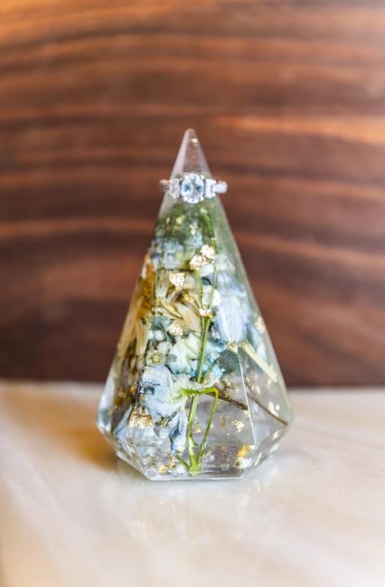May include: A clear, geometric ring holder with a silver ring on top. The holder is filled with dried blue and white flowers, gold flakes, and green stems. The ring has three clear stones.