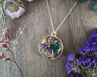 May include: A gold chain necklace with a round pendant containing pressed flowers, gold leaf, and green and purple stones.