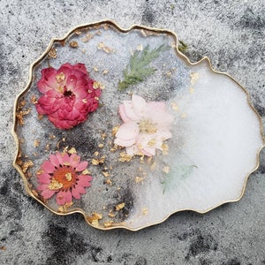May include: A white and gold resin coaster with a jagged edge, featuring three pressed flowers: a pink flower, a red flower, and a white flower with gold leaf accents.