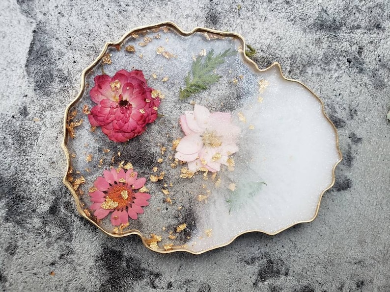 May include: A white and gold resin coaster with a jagged edge, featuring three pressed flowers: a pink flower, a red flower, and a white flower with gold leaf accents.