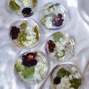 May include: Six clear resin coasters with dried flowers and gold leaf accents. Each coaster features a unique arrangement of white and purple flowers, green leaves, and gold flakes, creating a decorative and functional item.