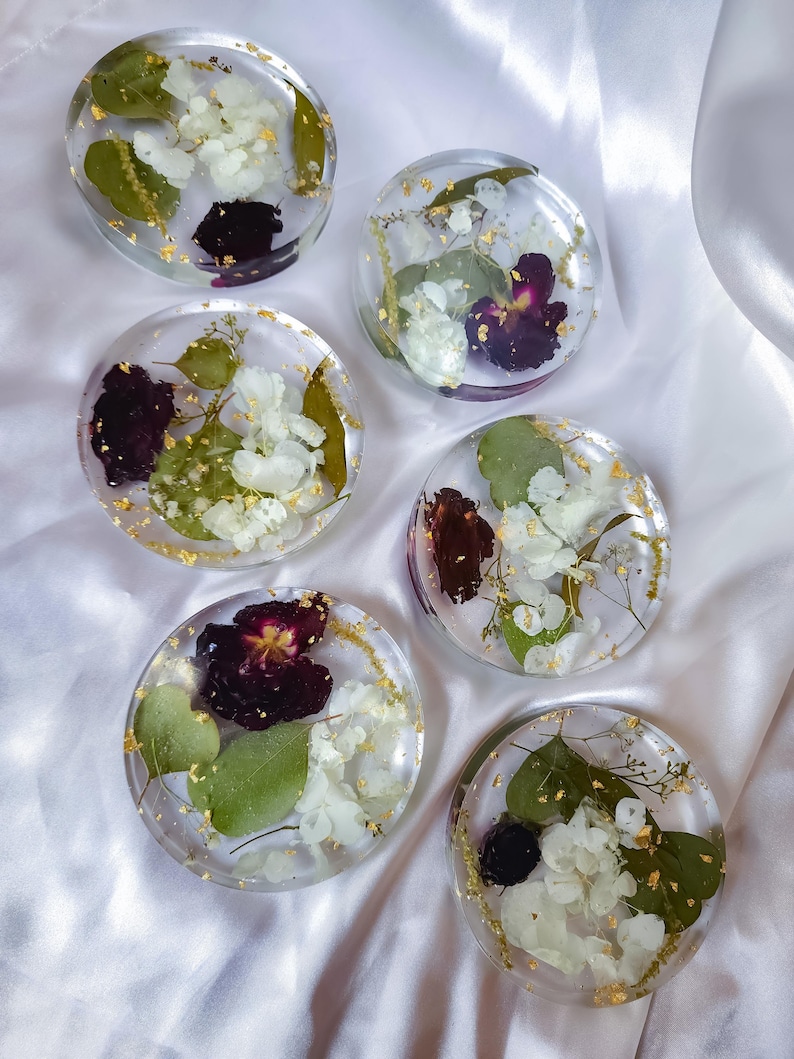 May include: Six clear resin coasters with dried flowers and gold leaf accents. Each coaster features a unique arrangement of white and purple flowers, green leaves, and gold flakes, creating a decorative and functional item.