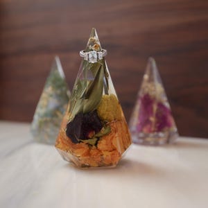 May include: A clear, faceted resin ring holder with dried flowers and a silver ring. The cone-shaped holder is filled with orange, yellow, and green dried flowers. Two other resin cone-shaped holders are in the background.