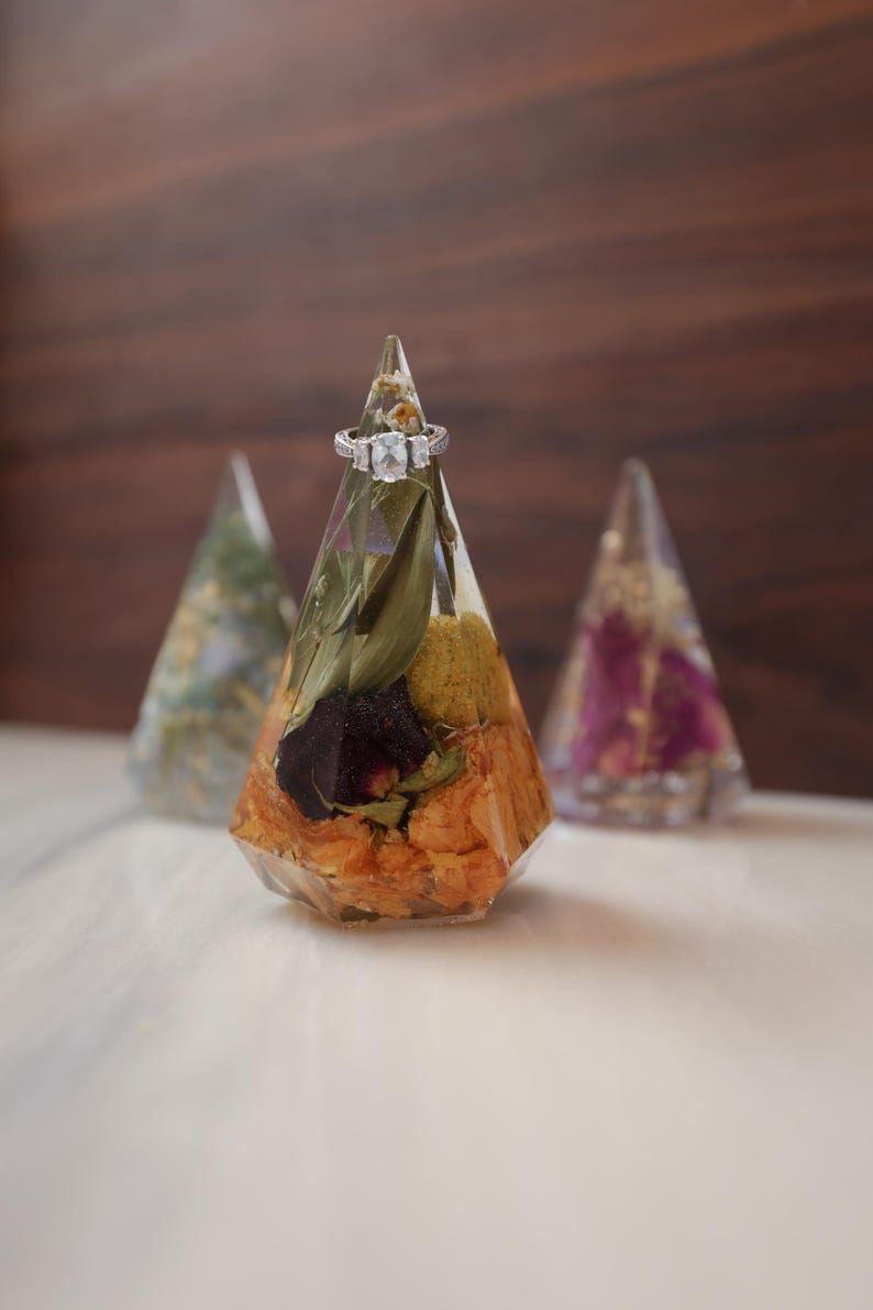 May include: A clear, faceted resin ring holder with dried flowers and a silver ring. The cone-shaped holder is filled with orange, yellow, and green dried flowers. Two other resin cone-shaped holders are in the background.