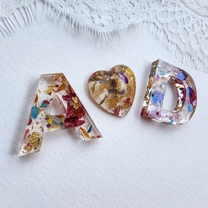 May include: Three resin ornaments, a heart and the letters A and D, filled with dried flowers and gold flakes.