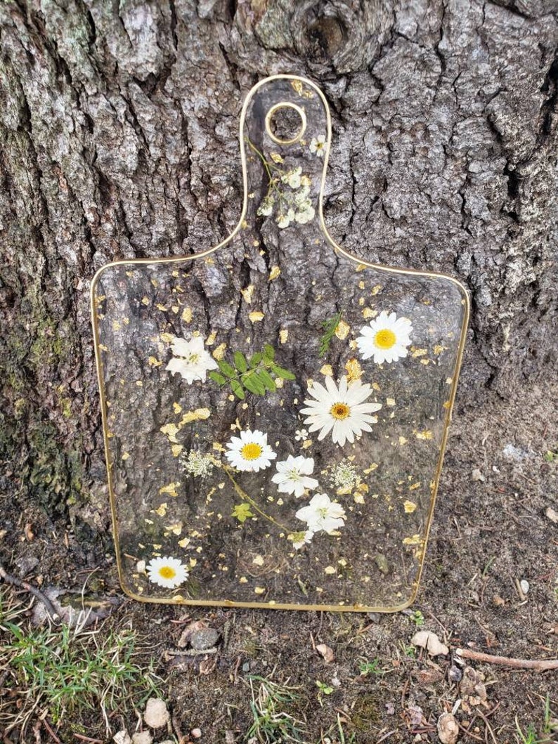Op de afbeelding: Een rechthoekig, transparant snijplank met een gouden rand en een gouden handvat. De plank is versierd met geperste witte bloemen en bladgoud.