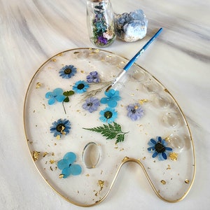 May include: A clear resin artist's palette with a gold rim, featuring pressed blue and white flowers, gold leaf, and a small glass vial filled with dried flowers.
