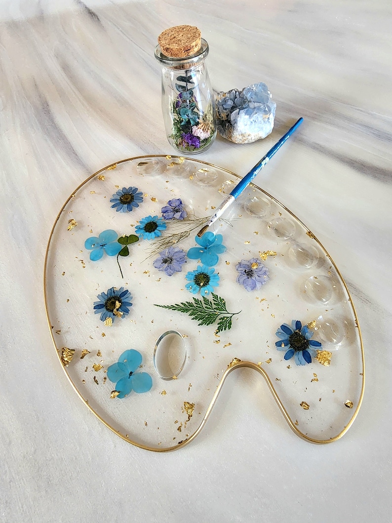 May include: A clear resin artist's palette with a gold rim, featuring pressed blue and white flowers, gold leaf, and a small glass vial filled with dried flowers.