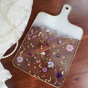Op de afbeelding: Een rechthoekige serveerschaal van wit en helder glas met een gouden rand en een handvat. De schaal is versierd met geperste paarse en witte bloemen en gouden vlokken.