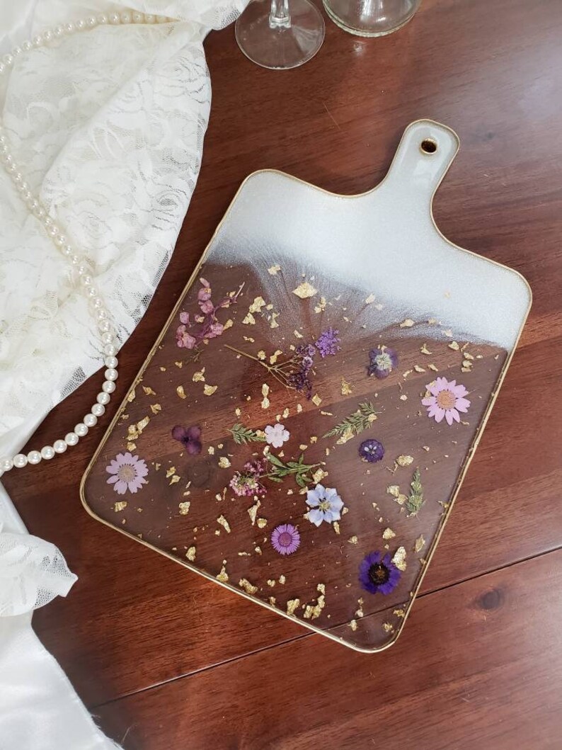 Op de afbeelding: Een rechthoekige serveerschaal van wit en helder glas met een gouden rand en een handvat. De schaal is versierd met geperste paarse en witte bloemen en gouden vlokken.