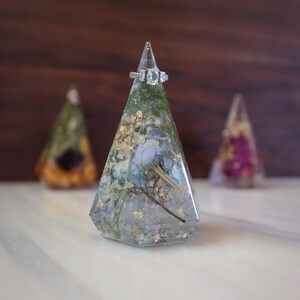 May include: A clear resin ring holder with dried flowers and gold leaf inclusions. A silver ring with three stones sits atop the holder. Two other resin holders are slightly out of focus in the background.