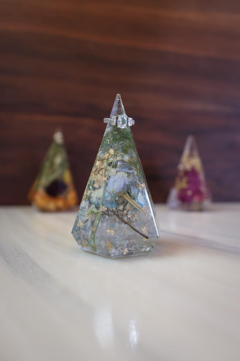 May include: A clear resin ring holder with dried flowers and gold leaf inclusions. A silver ring with three stones sits atop the holder. Two other resin holders are slightly out of focus in the background.