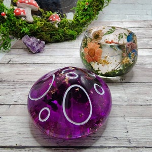 May include: Two resin crafts, one a purple dome with white circles and gold flecks, the other a clear dome with dried flowers and gold flecks.