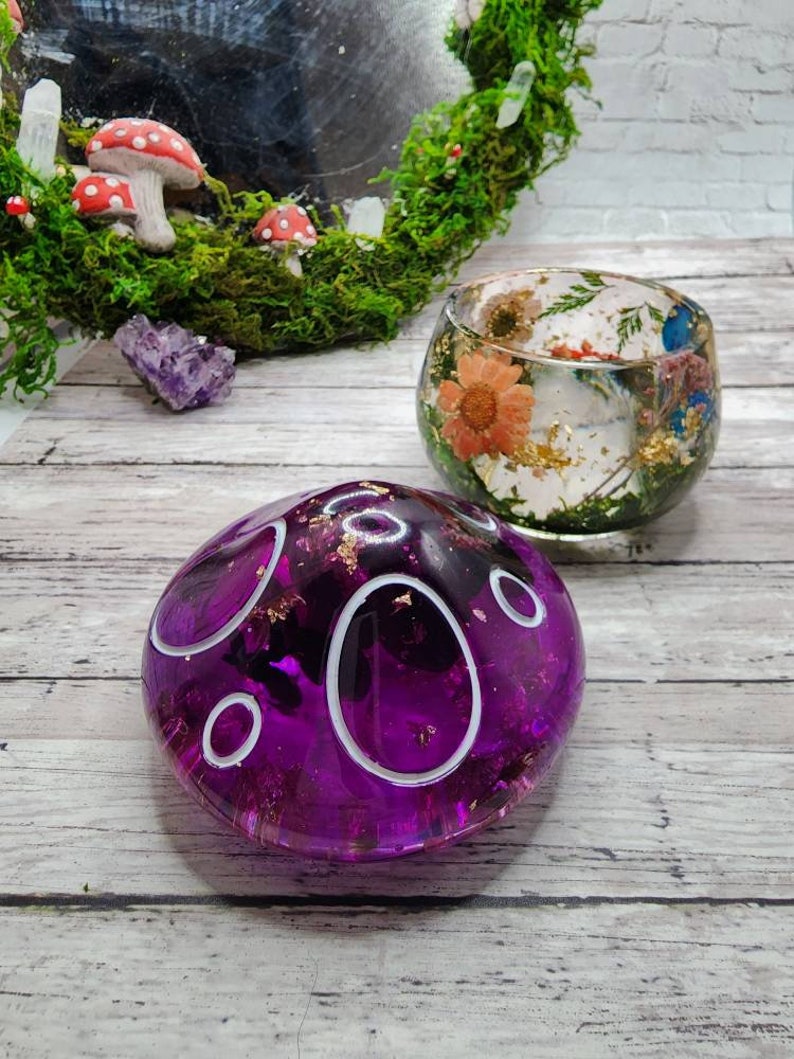 May include: Two resin crafts, one a purple dome with white circles and gold flecks, the other a clear dome with dried flowers and gold flecks.