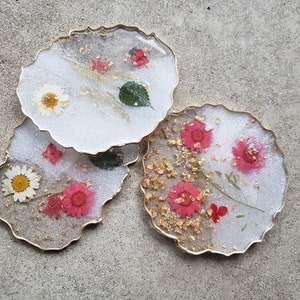 May include: Set of three white resin coasters with gold edges. Each coaster features pressed flowers and gold leaf accents. The flowers are pink, white, and red.