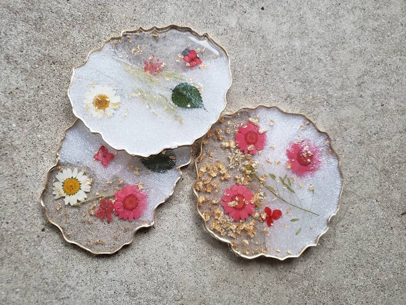 May include: Set of three white resin coasters with gold edges. Each coaster features pressed flowers and gold leaf accents. The flowers are pink, white, and red.