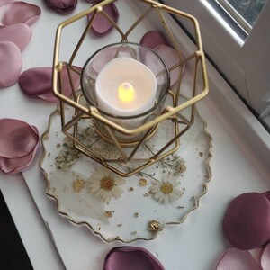 May include: A gold metal candle holder with a geometric design sits on a white and gold resin coaster with dried flowers and gold flakes. The candle holder is holding a clear glass candle with a warm white flame.