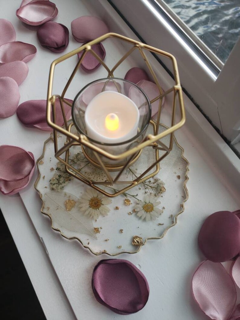 May include: A gold metal candle holder with a geometric design sits on a white and gold resin coaster with dried flowers and gold flakes. The candle holder is holding a clear glass candle with a warm white flame.