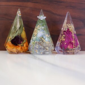 May include: Three clear resin ring holders in a triangular shape, each containing dried flowers and gold flakes. One holder displays a diamond ring. The holders are set against a wooden background.