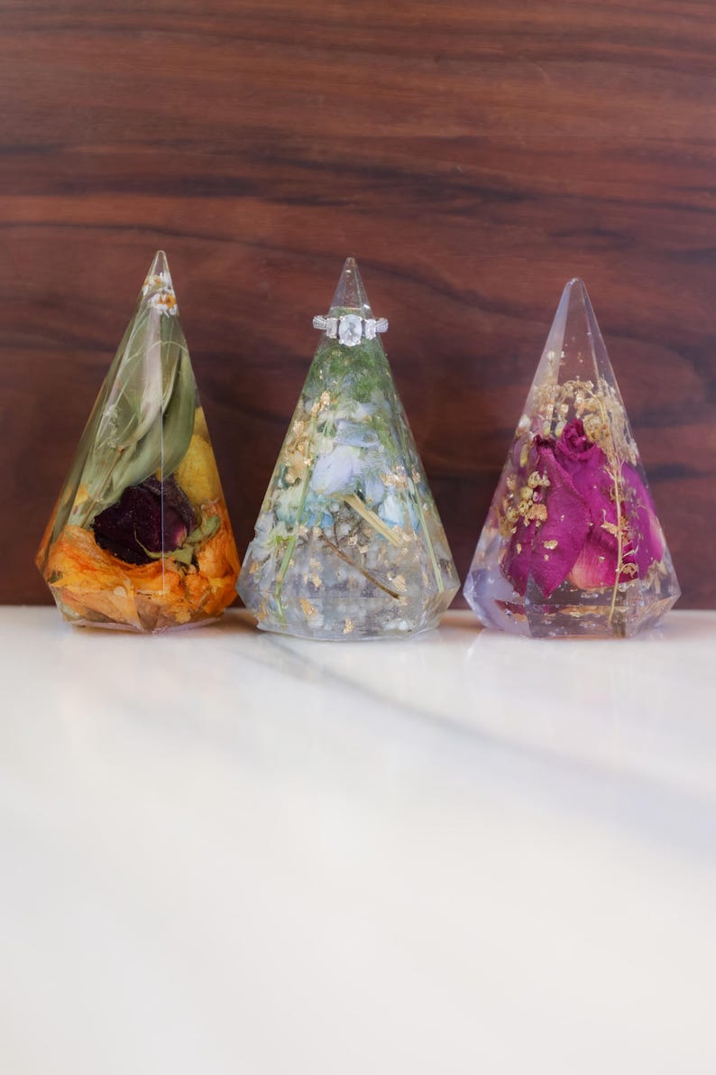 May include: Three clear resin ring holders in a triangular shape, each containing dried flowers and gold flakes. One holder displays a diamond ring. The holders are set against a wooden background.