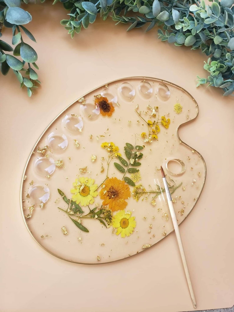 May include: A round, white, resin paint palette with a gold rim. The palette has 12 circular wells and is decorated with pressed flowers and gold leaf. A wooden paintbrush is lying next to the palette.