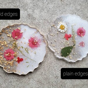 May include: Two round resin coasters with pressed flowers and gold leaf. The coaster on the left has a gold edge, while the coaster on the right has a plain edge. Both coasters are white with a clear resin top.