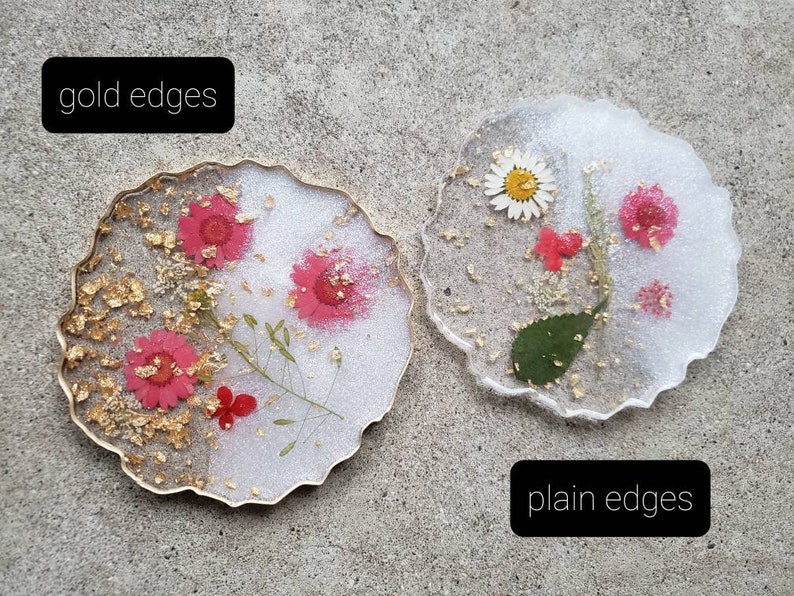 May include: Two round resin coasters with pressed flowers and gold leaf. The coaster on the left has a gold edge, while the coaster on the right has a plain edge. Both coasters are white with a clear resin top.
