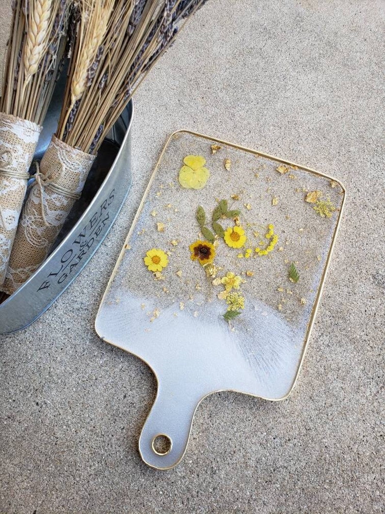 Op de afbeelding: Een witte serveerschaal met een gouden rand en handvat. De schaal is versierd met geperste bloemen en bladgoud. De schaal staat op een grijs betonnen oppervlak.