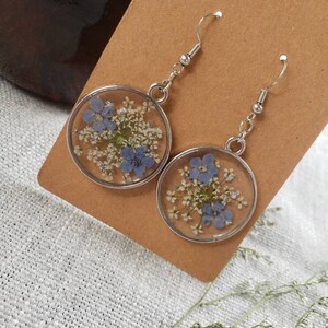 May include: A pair of silver earrings with a round, clear resin design featuring pressed blue and white flowers.