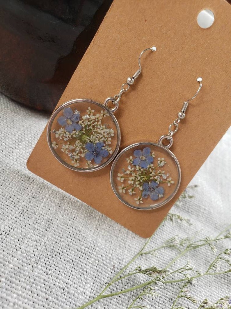 May include: A pair of silver earrings with a round, clear resin design featuring pressed blue and white flowers.