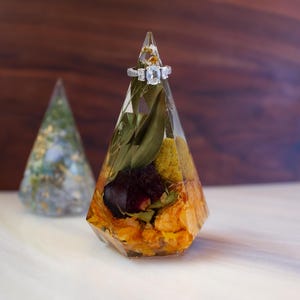 May include: A clear, geometric ring holder with dried flowers and a silver ring with three stones. The holder is filled with orange, yellow, and green dried flowers and leaves. A second, similar holder is in the background.