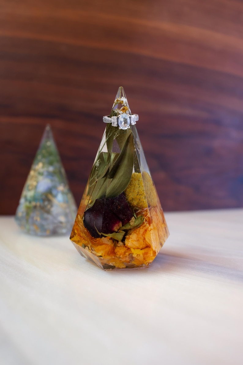 May include: A clear, geometric ring holder with dried flowers and a silver ring with three stones. The holder is filled with orange, yellow, and green dried flowers and leaves. A second, similar holder is in the background.
