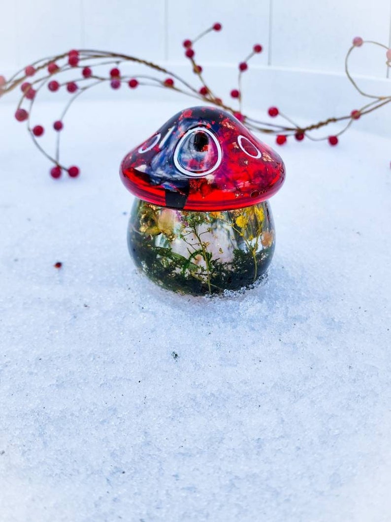 May include: A red and black mushroom-shaped glass ornament with a clear base filled with dried flowers and greenery. The ornament is sitting on a white surface.