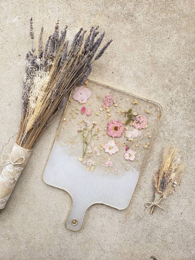 Op de afbeelding: Een wit serveerblad met een gouden rand en handvat, versierd met geperste roze en witte bloemen en gouden glitter. Het blad ligt op een lichtbruin oppervlak met gedroogde lavendel- en tarweboeketten.