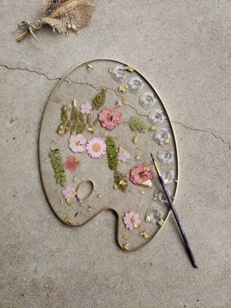 May include: A gold-rimmed, white ceramic palette with pressed flowers and gold leaf accents. The palette is on a grey concrete surface with a paintbrush lying next to it.